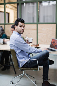 Portrait of businessman having coffee while using laptop at table in office