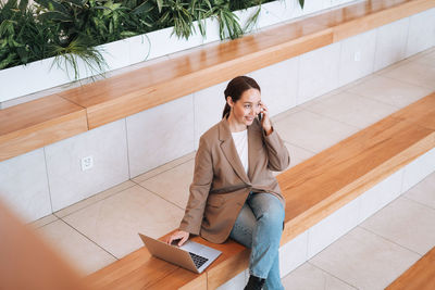 Adult smiling brunette business woman in beige suit and jeans working on laptop at public place