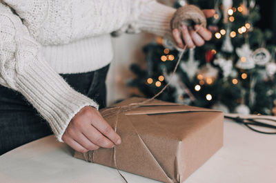 Midsection of woman wrapping christmas present in kraft paper in front of christmas tree at home