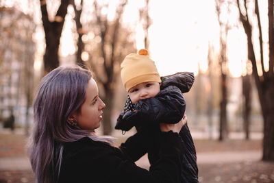 Young stylish mother holding cute baby son in park in autumn