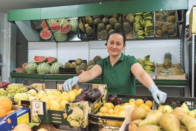 A shopkeeper is in front of her fruit shop looking at the camera