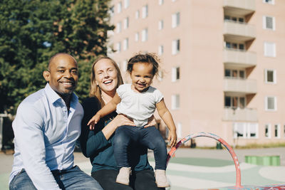 Portrait of happy multi-ethnic parents with daughter sitting at park in city