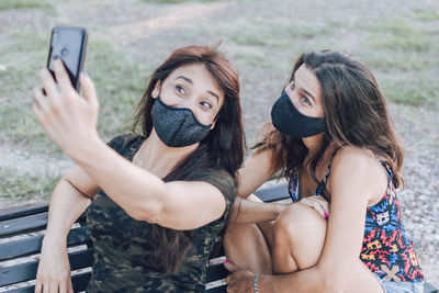 Women wearing mask doing selfie while sitting on bench