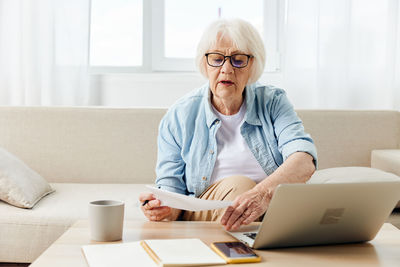 Senior woman holding paper sitting with laptop at home