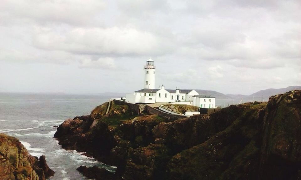 lighthouse, guidance, built structure, direction, sea, architecture, protection, water, safety, building exterior, cliff, sky, scenics, mountain, coastline, cloud - sky, nature, tower, tranquil scene, tranquility, beauty in nature, outdoors, cloudy, rock formation, day, non-urban scene, waterfront, lush foliage, distant, rocky coastline