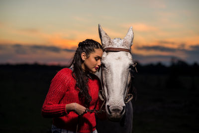 Young woman standing with white horse on field against sky during sunset