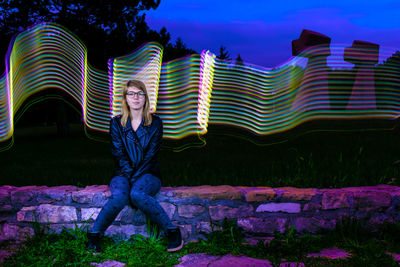 Full length of woman sitting against illuminated wall at night