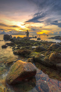 People on rocks by sea against sky during sunset