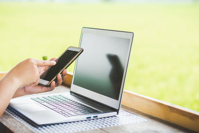 Cropped hands of woman using mobile phone by laptop on wooden table against landscape