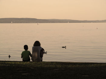 Rear view of mother with son sitting on lakeshore against sky during sunset