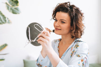 Young brunette woman in blue pajamas applies makeup in front of mirror sitting on bed at home