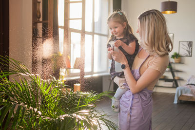 Young attractive woman with her toddler girl watering plants in home. making domestic work