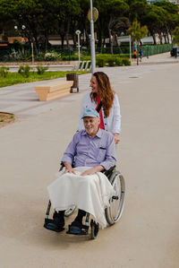 Smiling adult woman and senior man in wheelchair relaxing together in city on sunny day