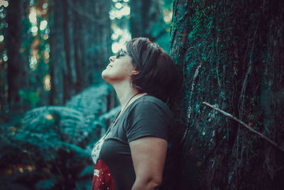 Side view of woman standing by tree trunk in forest