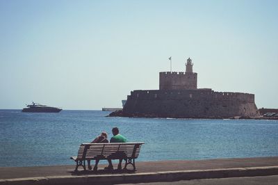 Rear view of man and woman sitting on bench on promenade with fort agios nikolaos in sea