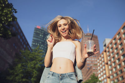 Portrait of happy woman with drink standing against buildings in city