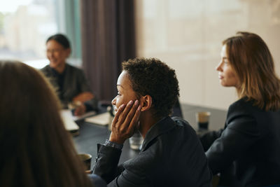 High angle view of short haired businesswoman leaning on elbow while sitting with colleagues in board room at office