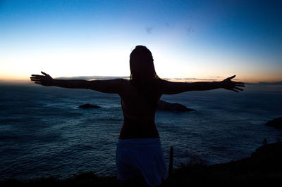Woman with arms outstretched standing by sea against sky during sunset