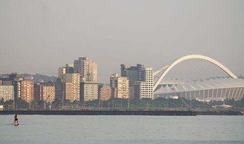 Man paddleboarding in sea by moses mabhida stadium in city against clear sky