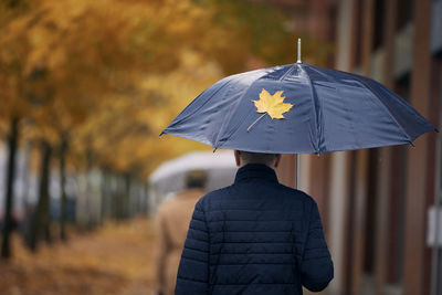 Man with umbrella walking under yellow maple trees on city street during autumn rainy day.