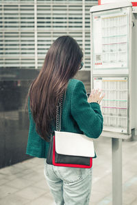 A young girl stands with her back near a bus stop with a timetable.