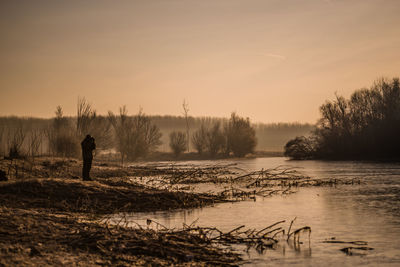 Man standing by lake against sky during sunset