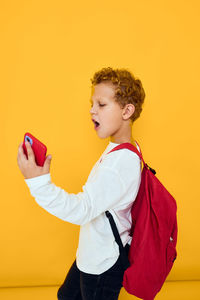 Side view of young woman using mobile phone while standing against yellow background