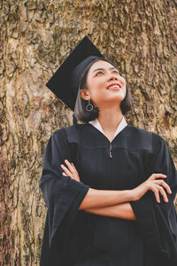 Thoughtful young woman in graduation gown with arms crossed standing by tree at park