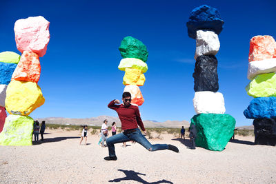 Full length of man jumping against colorful stacked rocks in desert during sunny day