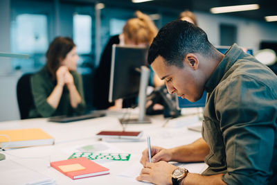 Side view of confident businessman writing on paper while female colleagues working at creative office