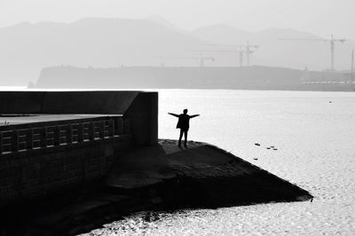 Silhouette of man with arms outstretched on pier at sea against sky