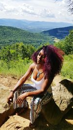 Woman with curly hair relaxing on rock against mountains during sunny day