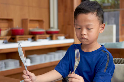Asian boy holds fork and knife prepare eating on table on buffet restaurant background