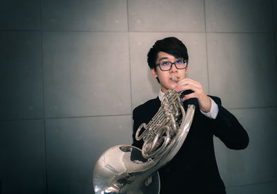 Portrait of teenage boy playing french horn while standing against wall