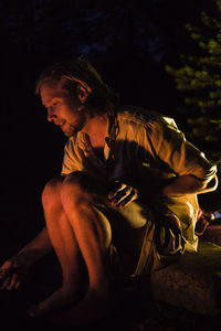 Side view of young man sitting by campfire