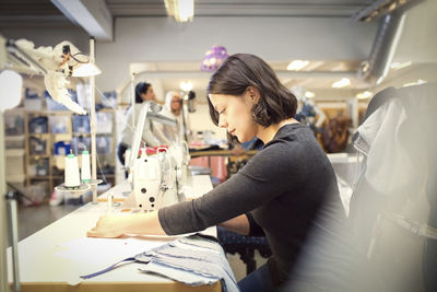 Side view of woman sewing fabric on machine while volunteers working in workshop