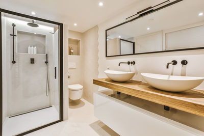 White sinks on wooden surface at wall with mirror placed in light bathroom with shower and tiles in apartment