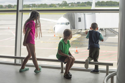 Girl with brothers watching preparation of airplane for boarding from airport terminal window