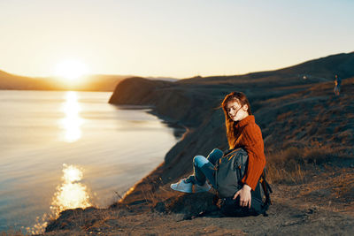Young woman sitting on rock against sky during sunset