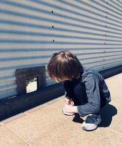 Boy looking at corrugated iron while crouching on sidewalk during sunny day