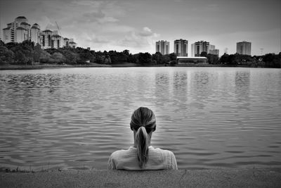 Rear view of woman sitting by lake against sky in city