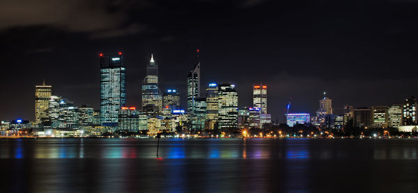 Illuminated buildings by river against sky at night