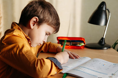 Cute 7 years old child doing his homework sitting by desk. boy writing in notebook. side view