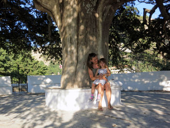 Smiling woman with daughter sitting by tree in park