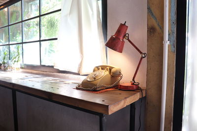 An old rotary phone and an electric lamp on a wooden table near the glass window and fabric curtain