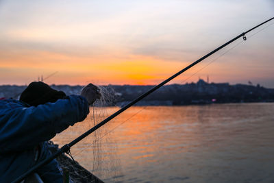 Rear view of man fishing in sea against sky during sunset