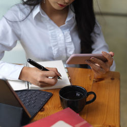 Midsection of woman using phone while writing on book at office