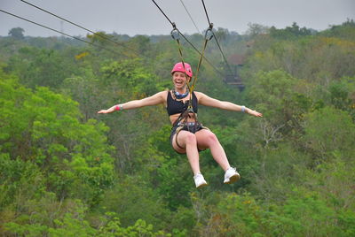 Full length of happy woman with arms outstretched zip lining against trees