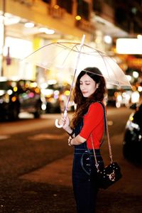 Side view of young woman holding umbrella on road in city at night