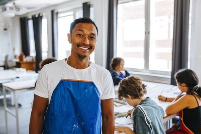 Portrait of smiling male art teacher wearing blue apron in classroom at school